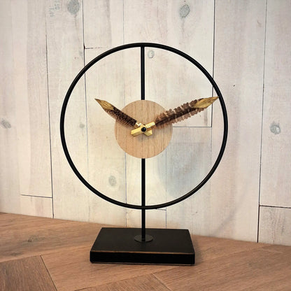 une horloge design sur pied en métal noir et bois, avec des plumes de faisan sur les aiguilles et finition dorée sur l'extrémité des plumes